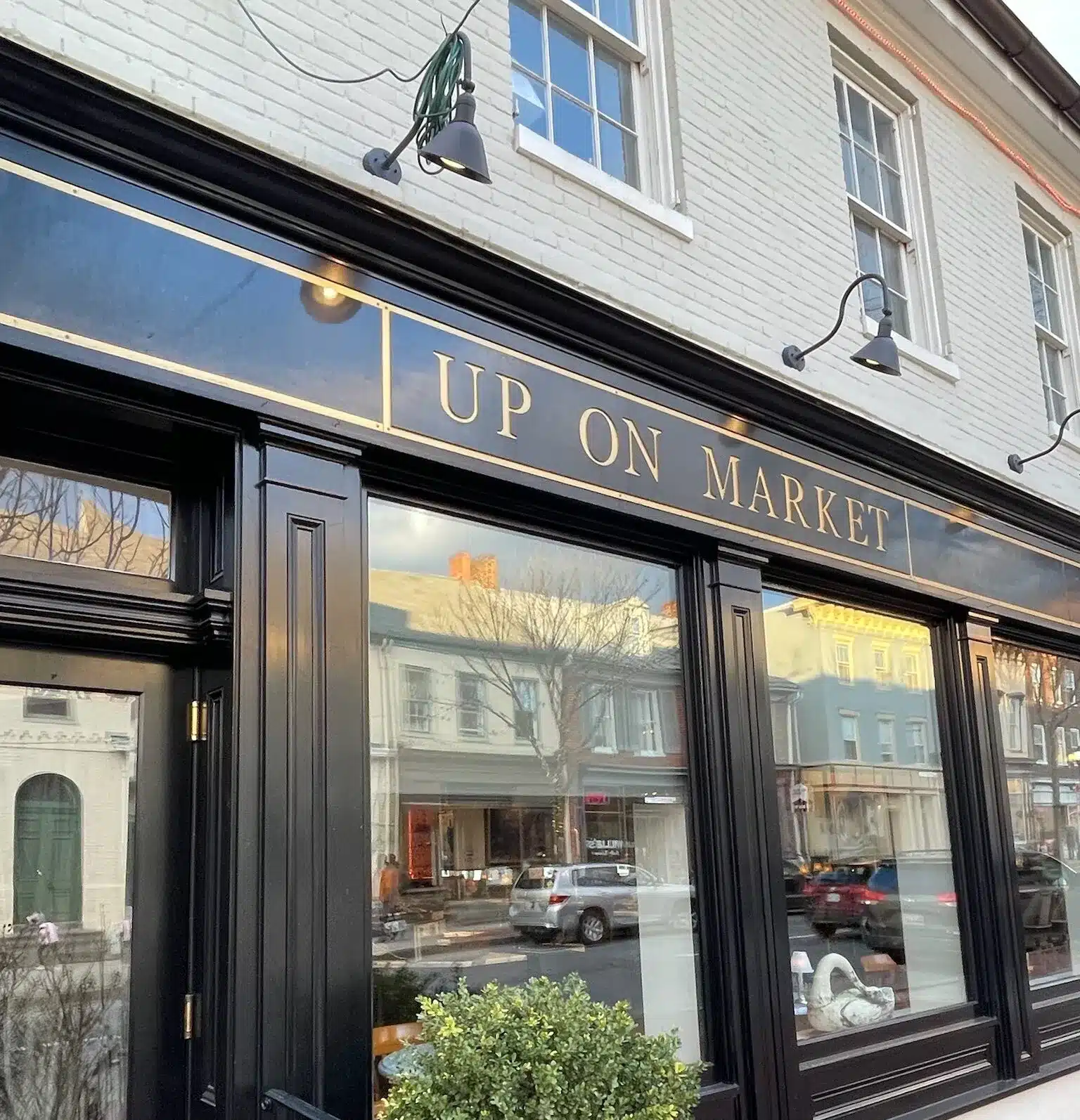 a Storefront with Black Trim and Large Windows Reflects Nearby Buildings and Cars Above a Gold Trimmed Black Sign Reads Up on Market One of the Best Bars in Frederick the White Brick Facade Features Potted Plants Outside