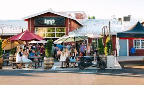 Upp Liquids Bend or a Lively Outdoor Patio at One of the Top Breweries in Bend Oregon with People Sitting at Picnic Tables Under Colorful Umbrellas Socializing the Red Brick Building Has a Sign That Reads Lpp String Lights Glow Above and a Scooter is Parked in Front