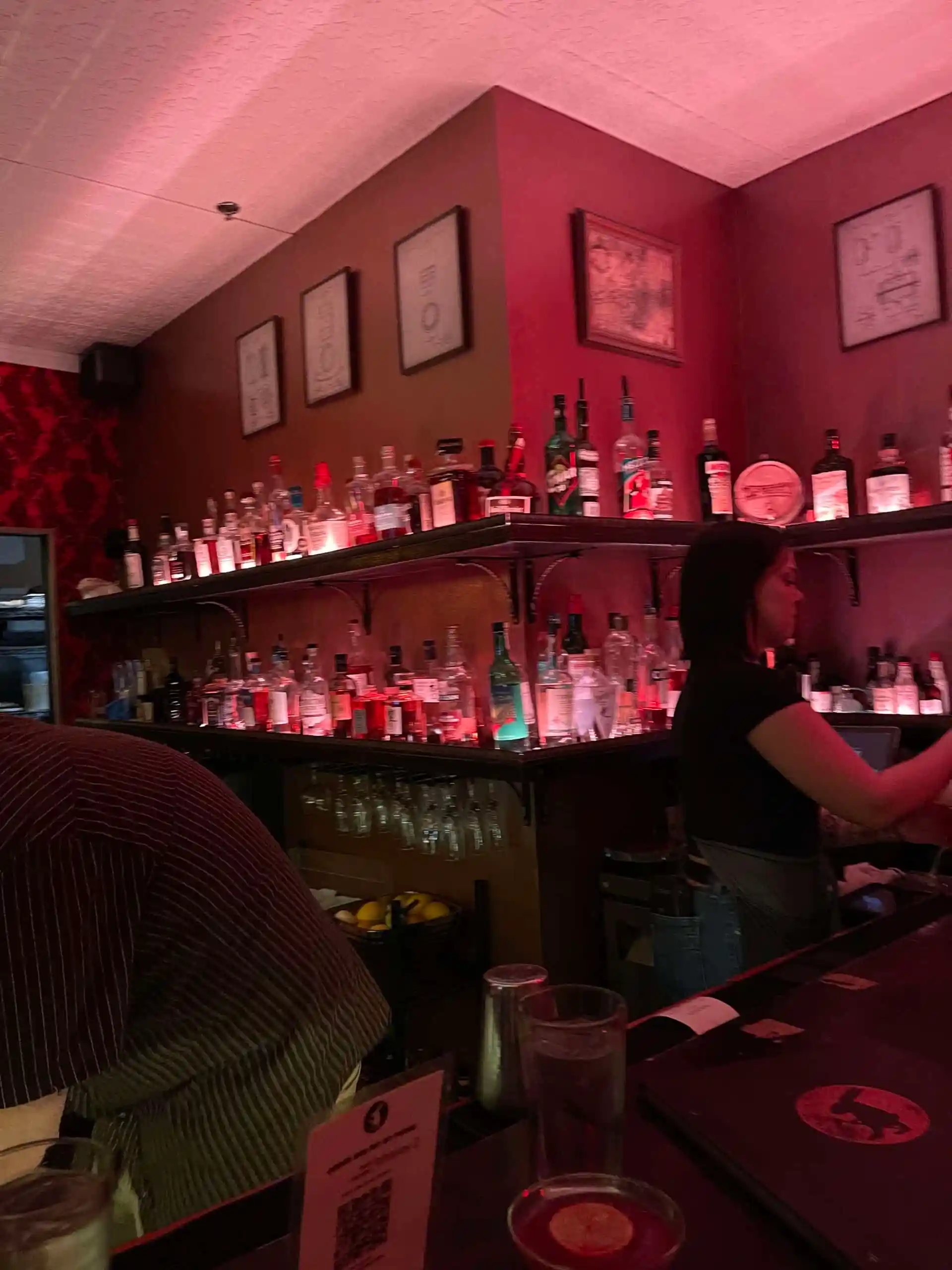 a Dimly Lit Bar with Red Walls Shelves Stocked with Bottles of Liquor and Framed Artjust the Kind of Cozy Spot Youd Find in Bend Oregon a Bartender Prepares Drinks As a Person in a Striped Shirt Sits at the Bar in the Foreground