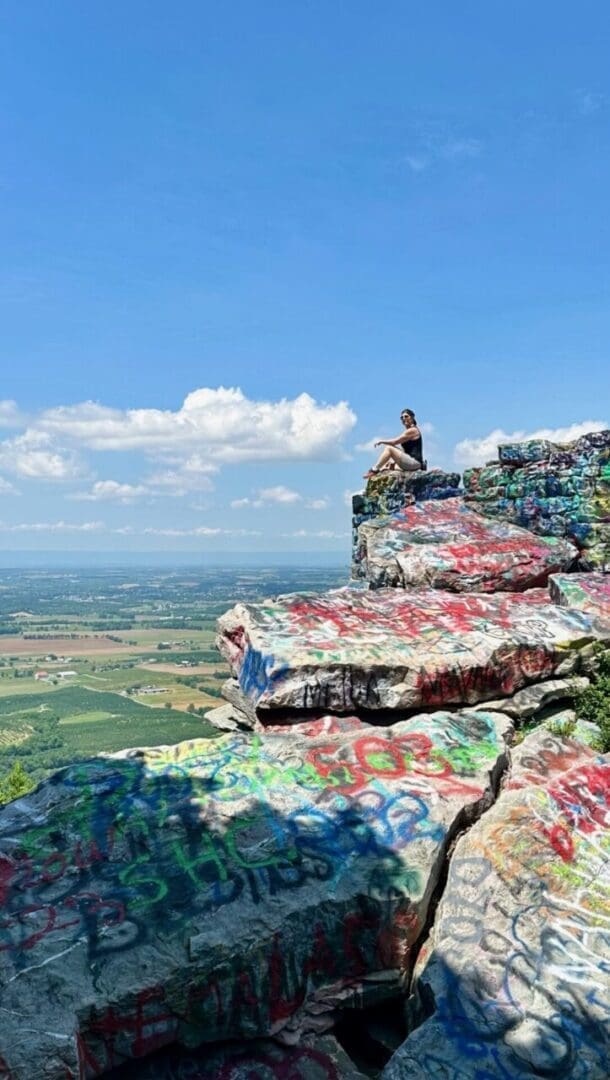 a Person Sits on the Edge of a Layered Rocky Cliff Covered in Colorful Graffitione of the Best Hikes Around Frederick Mdoverlooking a Wide Expanse of Farmland and Green Fields Under a Bright Blue Sky with Scattered Clouds