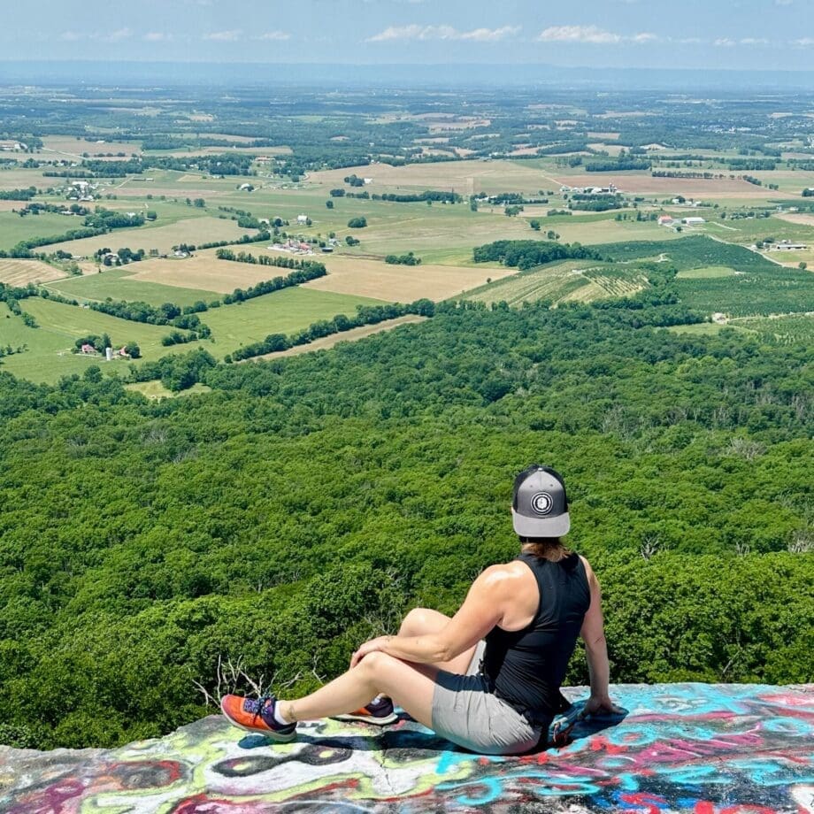 a Person in a Black Tank Top Gray Shorts and a Cap Sits on a Colorful Graffiti Covered Rockone of the Best Hikes Around Frederick Mdtaking in Sweeping Views of Farmland Fields Distant Hills and Lush Green Forest Under a Clear Sky