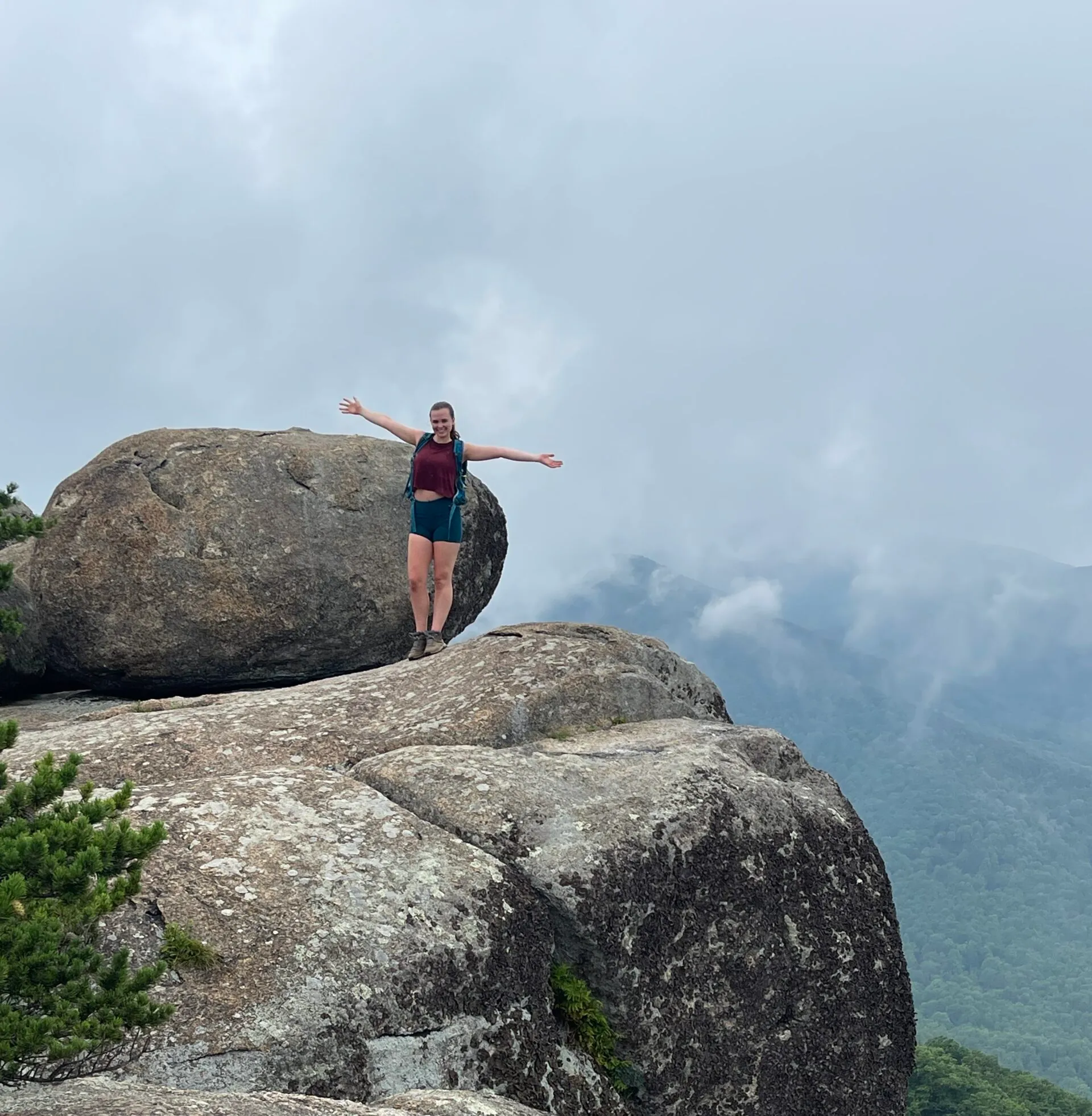 a Person with Arms Outstretched Stands on a Large Rocky Ledge Next to a Massive Boulderperhaps Discovering One of the Best Hikes Around Frederick Md They Wear a Red Shirt and Teal Shorts Framed by Misty Mountains and Forest in the Cloudy Background