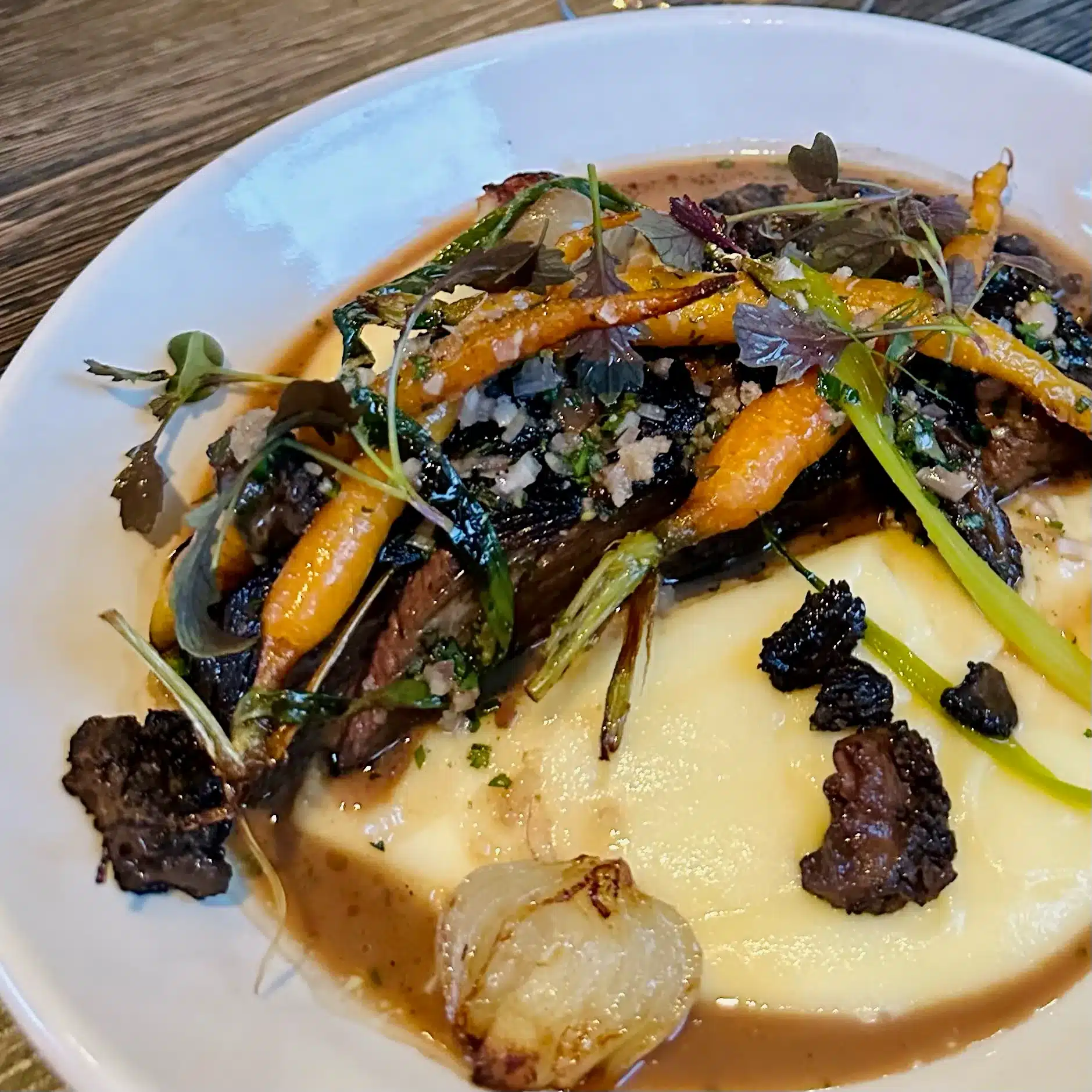 a White Plate Holds Creamy Mashed Potatoes Topped with Braised Beef Roasted Baby Carrots Morel Mushrooms and Fresh Herbs Inspired by Dishes Served at the Best Bars in Frederick Its Finished with Rich Brown Sauce and Microgreens on a Wooden Table