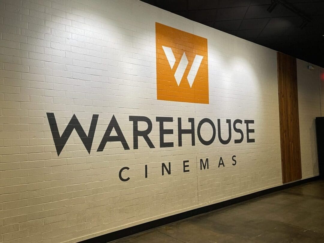 a White Brick Wall Displays the Warehouse Cinemas Logo in Bold Black Letters Above an Orange Square Features a Stylized White W This Frederick Movie Theatre Setting Includes Polished Concrete Floors and Partial Wood Paneling in an Indoor Corridor