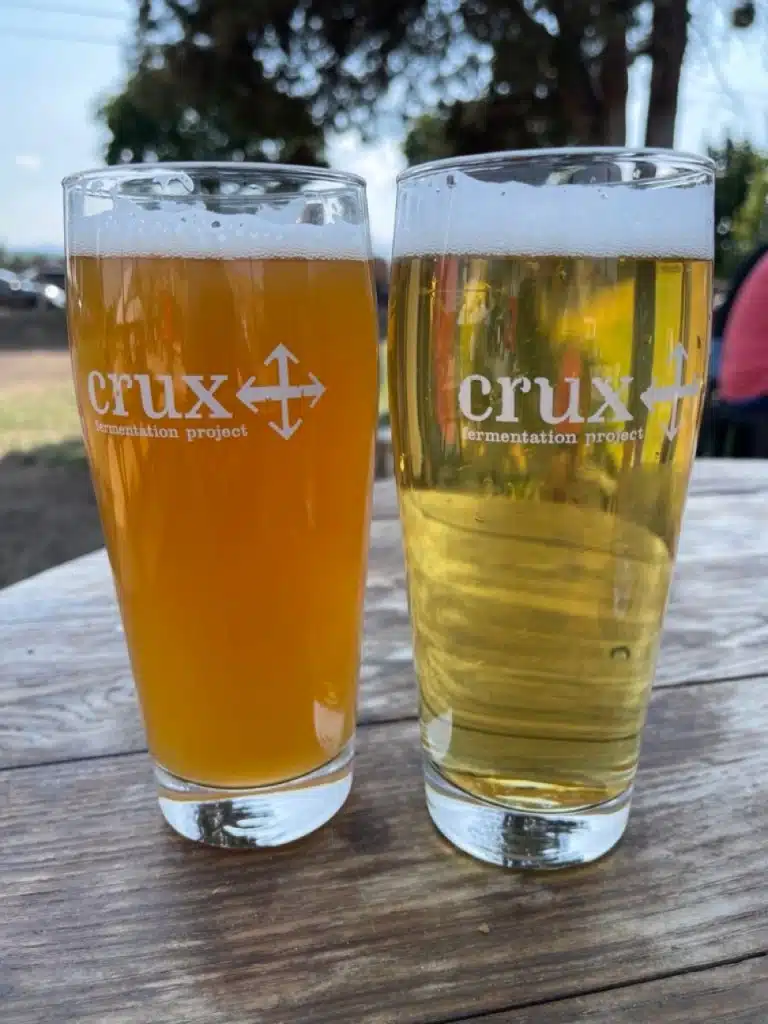 Two Pint Glasses with Crux Fermentation Project Logos Sit on a Wooden Table Outdoors in Bend Oregon One Holds a Hazy Beer the Other a Golden Brewperfect for Enjoying One of the Best Things to Do in Bend Relaxing with Local Craft Beer