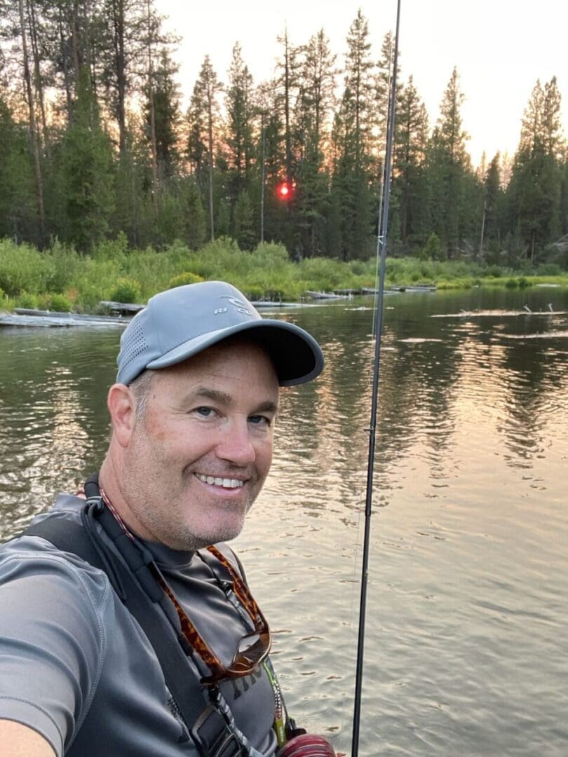 a Man in a Gray Shirt and Cap Smiles While Holding a Fishing Rod Standing in Shallow Water As the Sun Setsa Peaceful Outdoor Scene That Highlights One of the Top Things to Do in Bend or the Calm Water Reflects the Sky and Forest Around Him