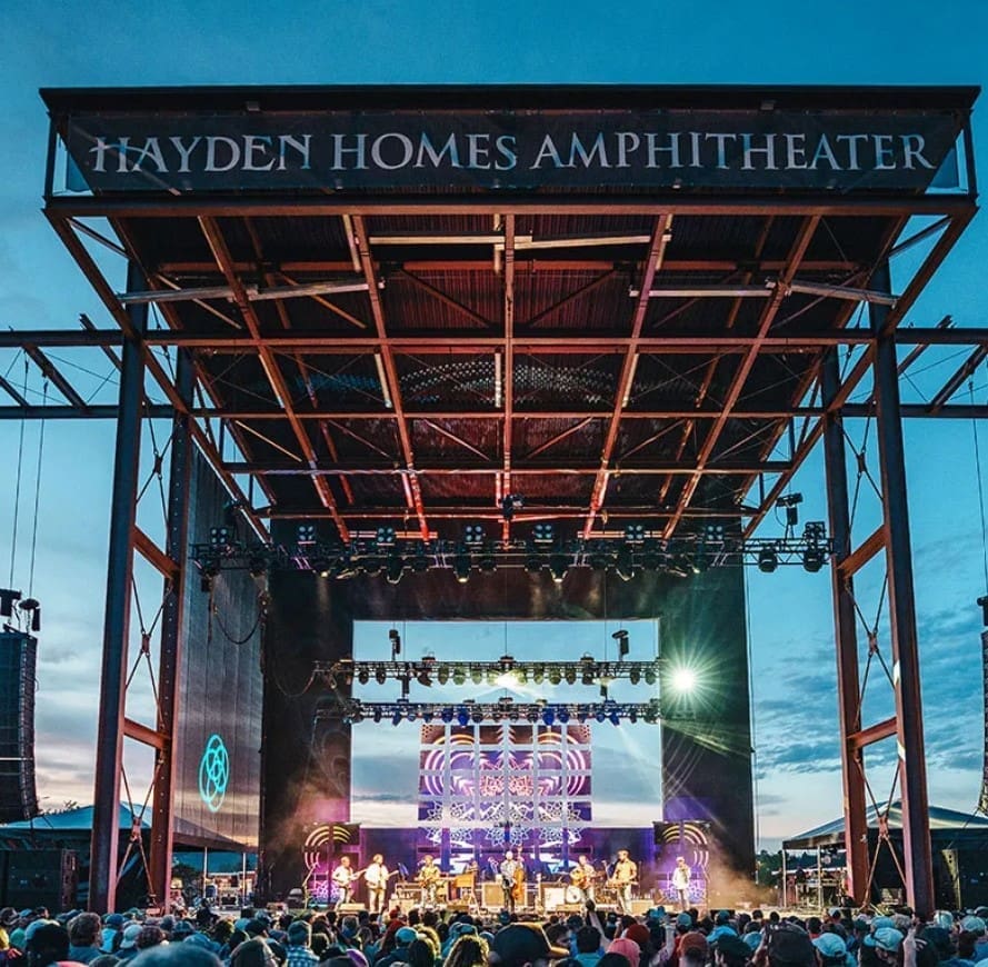 a Crowd Gathers at an Outdoor Concert in Front of a Large Stage Under a Metal Canopy Labeled Hayden Homes Amphitheaterone of the Top Things to Do in Bend or the Stage Glows with Colorful Lights As Musicians Perform into the Dusk