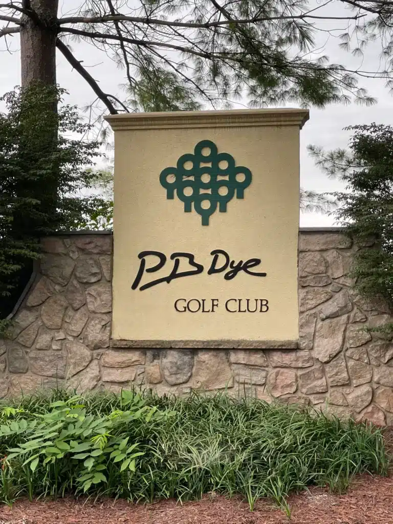 a Stone Wall Displays a Beige Sign with a Green Abstract Golf Ball Logo Above the Words Pb Dye Golf Club in Black Script Bushes Tall Grass and Trees Surround the Sign Near New Market Md the Sky is Overcast