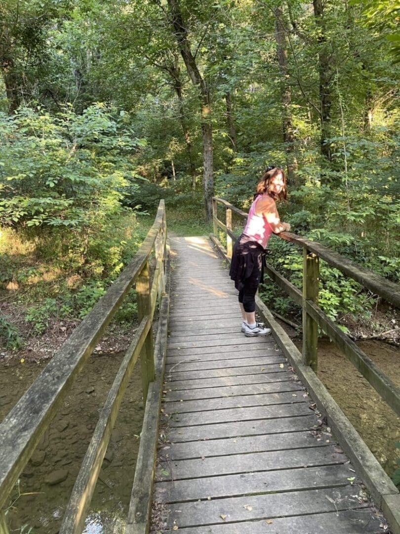 a Woman Stands on a Wooden Footbridge in a Lush Green Forest Along One of the Best Hikes Around Frederick Md She Leans on the Railing Sunlight Filtering Through the Trees and Casting Dappled Light on the Bridge and Surrounding Foliage
