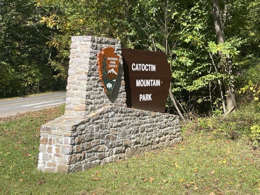 a Stone and Wood Sign at the Roadside Reads Catoctin Mountain Park with a National Park Service Emblem Green Trees and Grass Surround the Signan Inviting Gateway to Some of the Best Hikes Around Frederick Md As Sunlight Filters Through the Foliage
