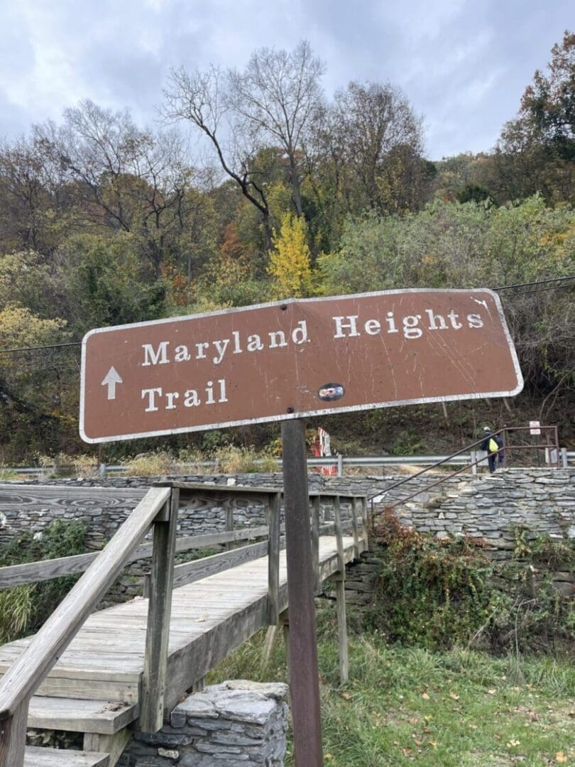 a Brown Wooden Sign Reads Maryland Heights Trail with a White Arrow Pointing Up the Sign Stands Beside a Wooden Footbridgeone of the Best Hikes Around Frederick Mdframed by Stone Walls and Autumn Trees Under a Cloudy Sky