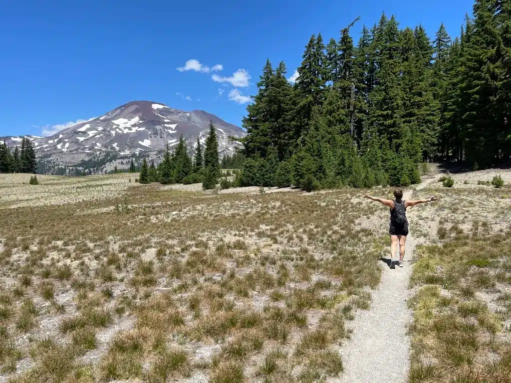 a Person Walks with Arms Outstretched on a Narrow Trail Through a Grassy Field Near Bend Oregon Toward a Snow Capped Mountain Surrounded by Tall Green Pine Trees Under a Bright Blue Sky