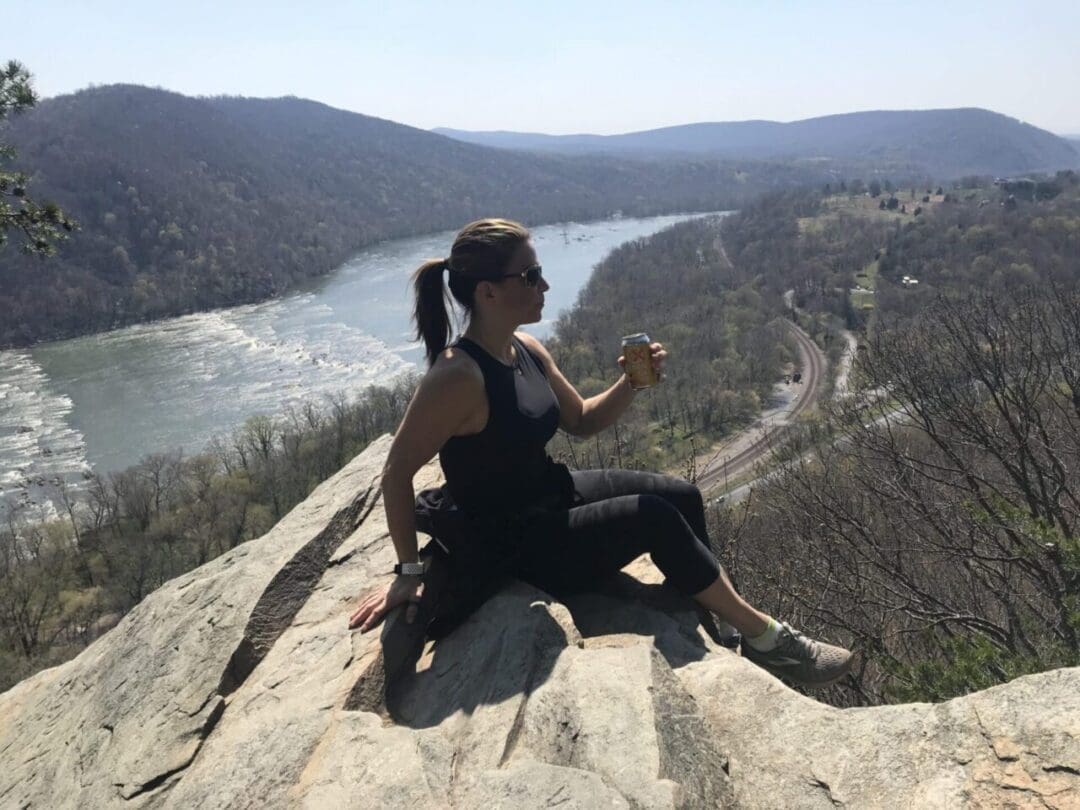 a Woman in Athletic Wear Sits on a Rocky Outcrop Overlooking a River and Forested Hills Enjoying One of the Best Hikes Around Frederick Md She Holds a Drink Can Wears Sunglasses and Gazes at Train Tracks Below Under a Hazy Sky