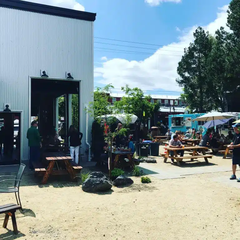 People Sit at Wooden Picnic Tables Outside a Metal Building on a Sunny Day Enjoying Food from Local Bend Restaurants Surrounded by Trees Rocks and Food Trucks the Area Has a Relaxed Social Atmosphere Under a Blue Sky with Clouds