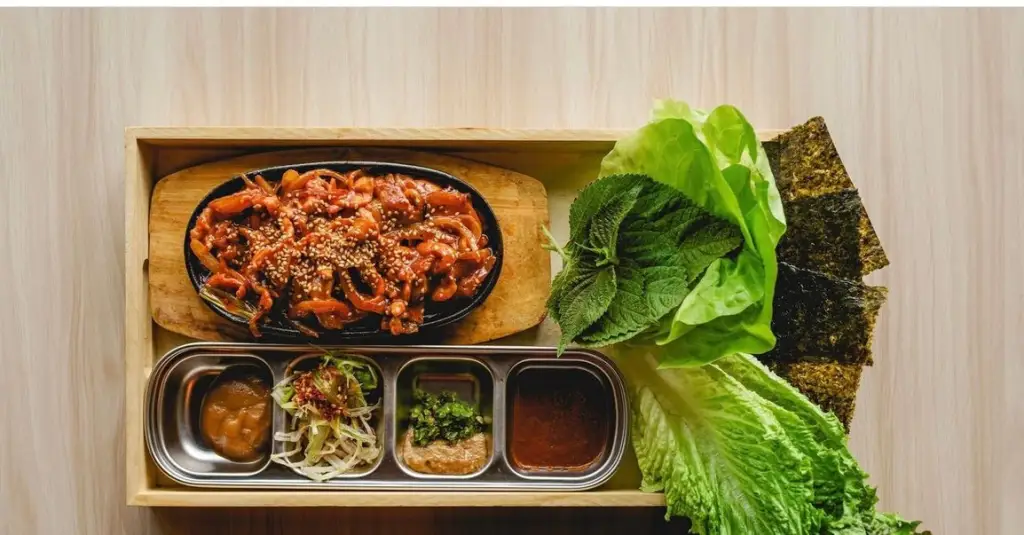 at Bend Restaurants a Wooden Tray Holds a Sizzling Dish of Stir Fried Meat Topped with Sesame Seeds Four Small Containers of Sauces and Vegetables Leafy Greens Shiso Leaves and Neatly Arranged Sheets of Dried Seaweed on the Side