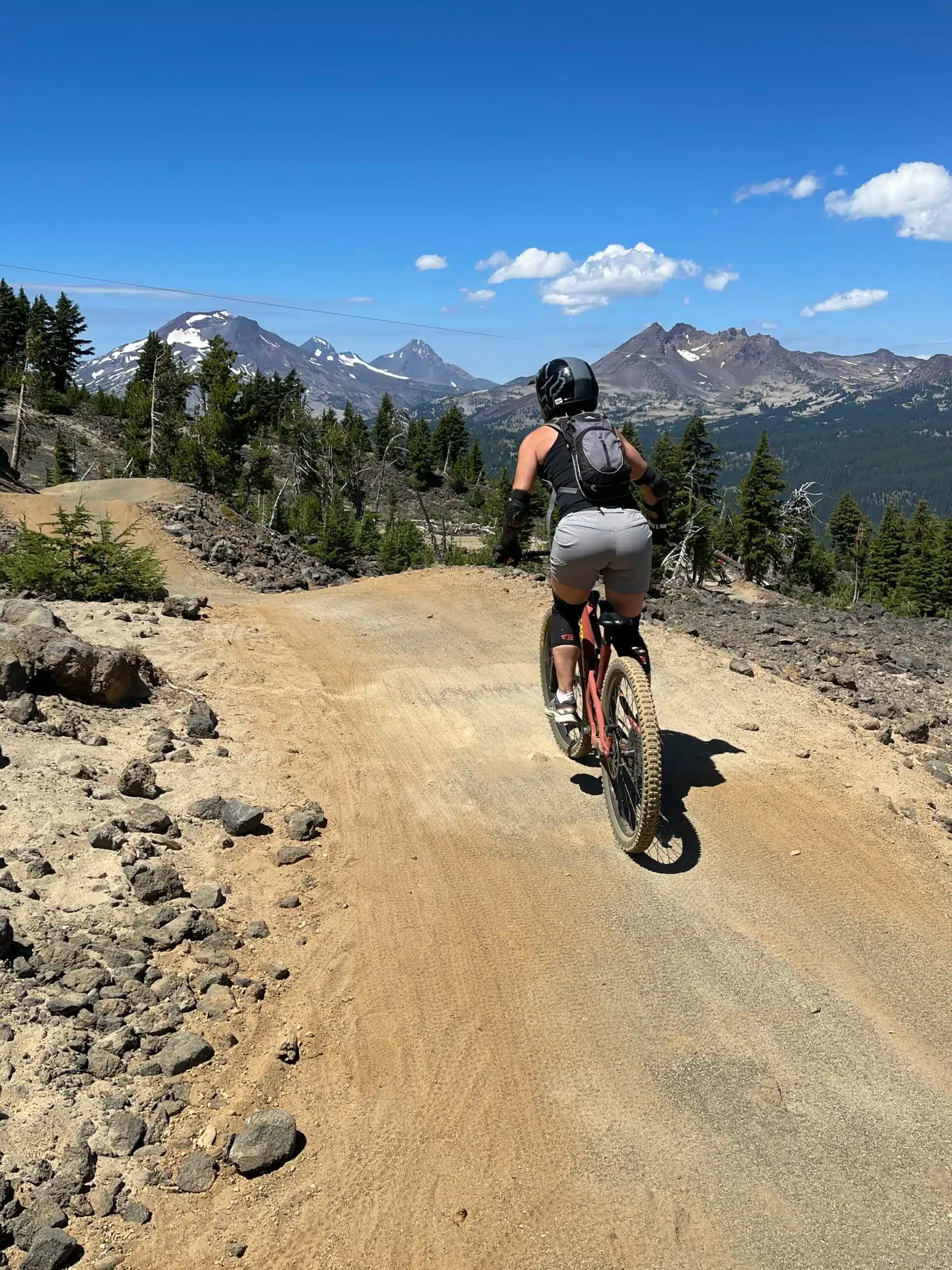 a Cyclist Wearing a Helmet Backpack and Shorts Rides a Mountain Bike Up a Dirt Trail Surrounded by Rocks and Pine Treesone of the Top Things to Do in Bend Orwith Snow Capped Mountains and a Clear Blue Sky in the Background