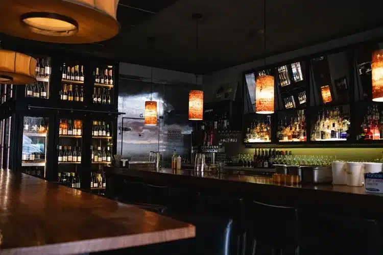 a Dimly Lit Bar with Wooden Countertops Hanging Amber Pendant Lights and Shelves Lined with Bottles of Wine and Liquor Barstools Line the Counter While Mirrors Reflect the Cozy Charm Found in the Best Bend Restaurants