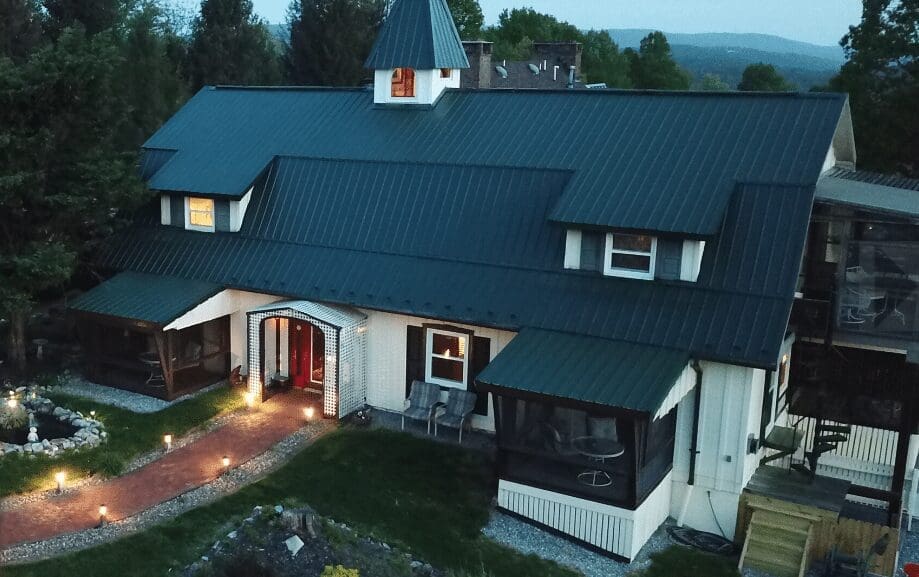 a Large Dark Roofed House with Dormer Windows and a Central Peaked Tower Resembling the Charm of Historic Hotels in Frederick Md It Features Screened Porches Garden Beds a Red Brick Walkway and Green Trees Under a Cloudy Sky
