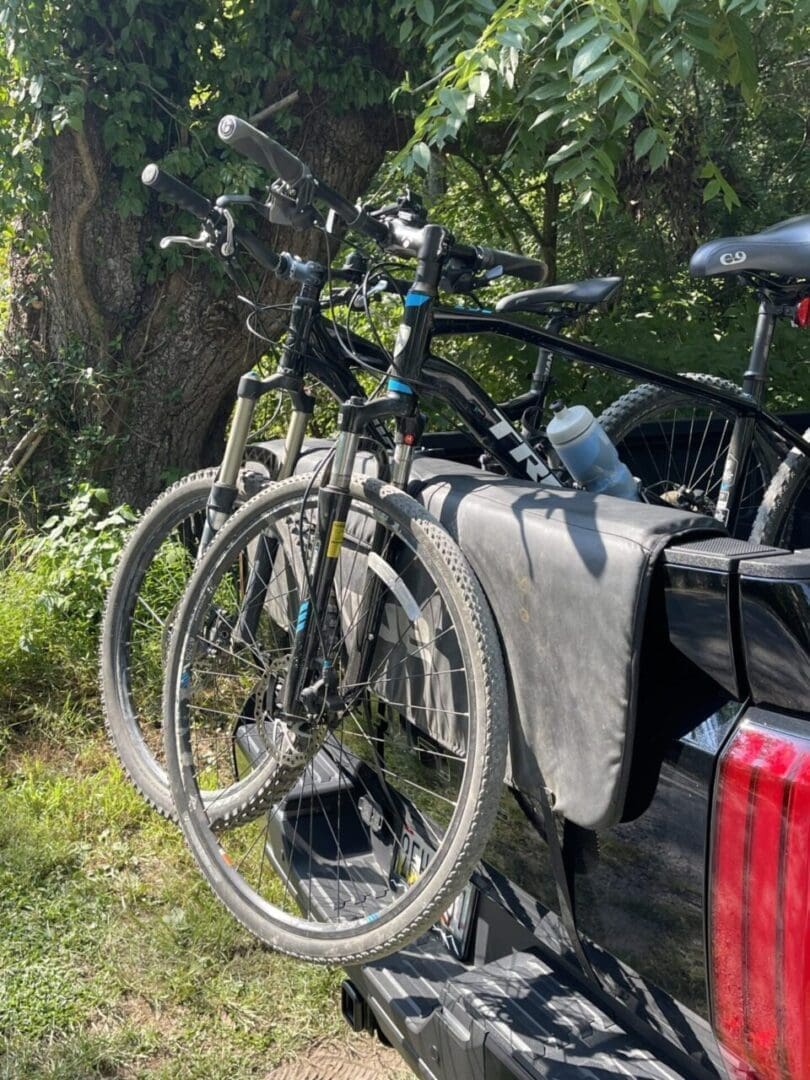 Two Black Mountain Bikes Are Mounted on the Tailgate of a Black Pickup Truck Secured by a Padded Tailgate Cover Outdoors Near Little Bennett Bike Trails Green Grass and Leafy Trees in the Background Suggest a Sunny Day