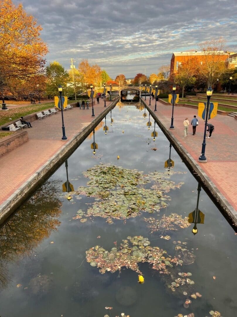 View of a Canal Lined with Lily Pads and Brick Walkways in Frederick Md People Stroll Under Autumn Trees and Yellow Banners Boutique Hotels Frederick Md Are Nearby with Buildings in the Background Beneath a Partly Cloudy Sky