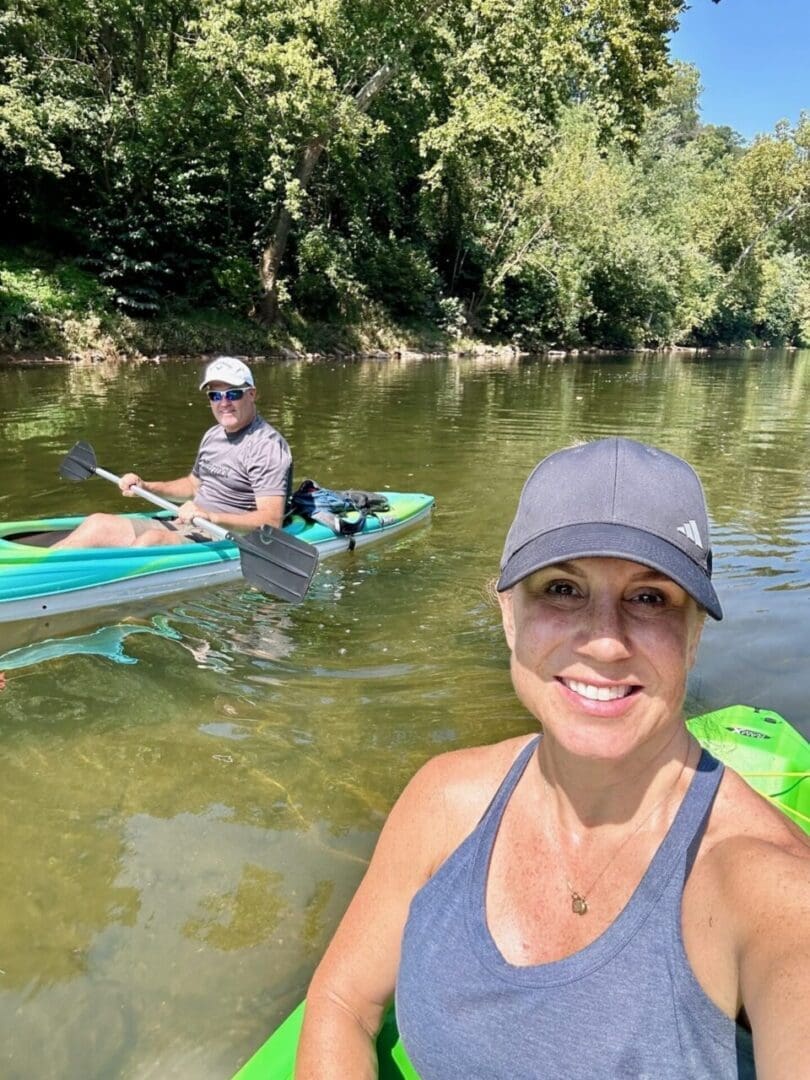 a Woman in a Blue Tank Top and Baseball Cap Smiles at the Camera While Kayaking the Monocacy River a Man in Sunglasses Paddles Behind Her As They Enjoy a Sunny Day Surrounded by Greeneryperfect for Kayak Monocacy River Adventures