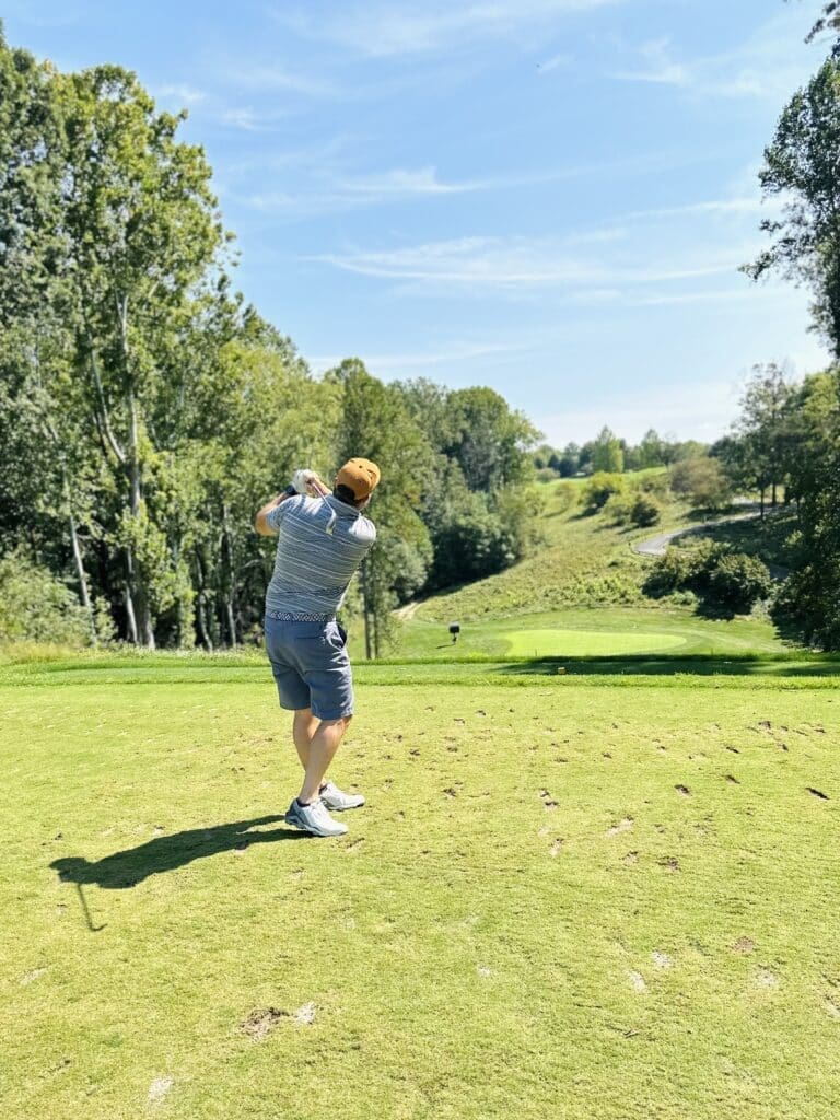 a Man in a Striped Shirt Shorts Sneakers and an Orange Cap Swings a Golf Club on a Green Course Surrounded by Tall Trees and Rolling Hills Enjoy Scenes Like This at Golf Courses Near Frederick Md Under the Clear Blue Sky