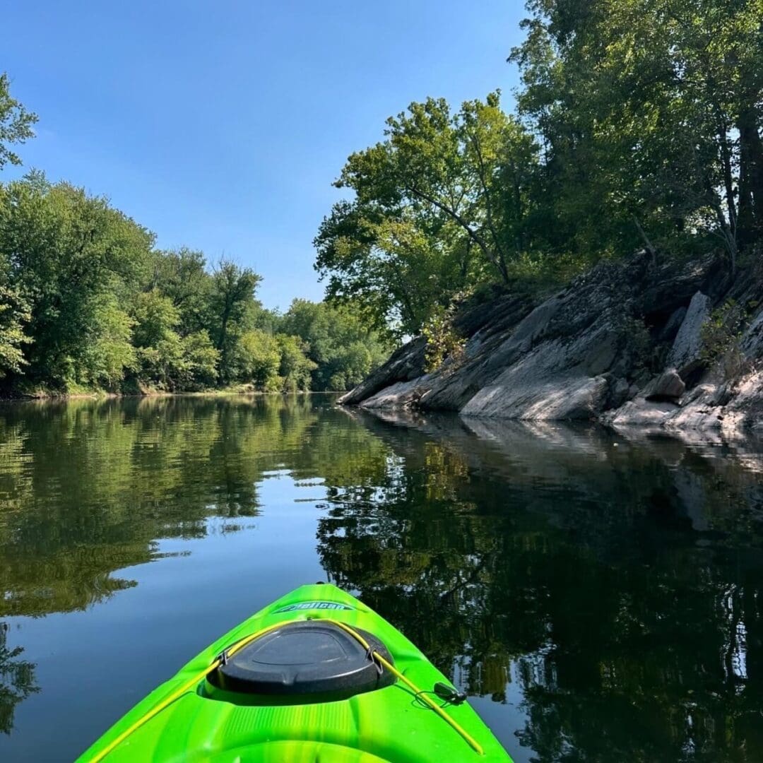 a Bright Green Kayak Glides Down the Calm Reflective Monocacy River Near Frederick Md Bordered by Lush Trees and a Rocky Shoreline Under a Clear Blue Sky Sunlight Casts Gentle Shadows on the Water and Rocks in This Peaceful Kayak Monocacy Experience