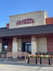 the Image Shows the Exterior of Outback Steakhouse Known As the Best Steakhouse Frederick Md the Building Features Tan Stone Walls a Red and White Sign and Flower Planters with White and Red Blooms on a Clear Sunny Day