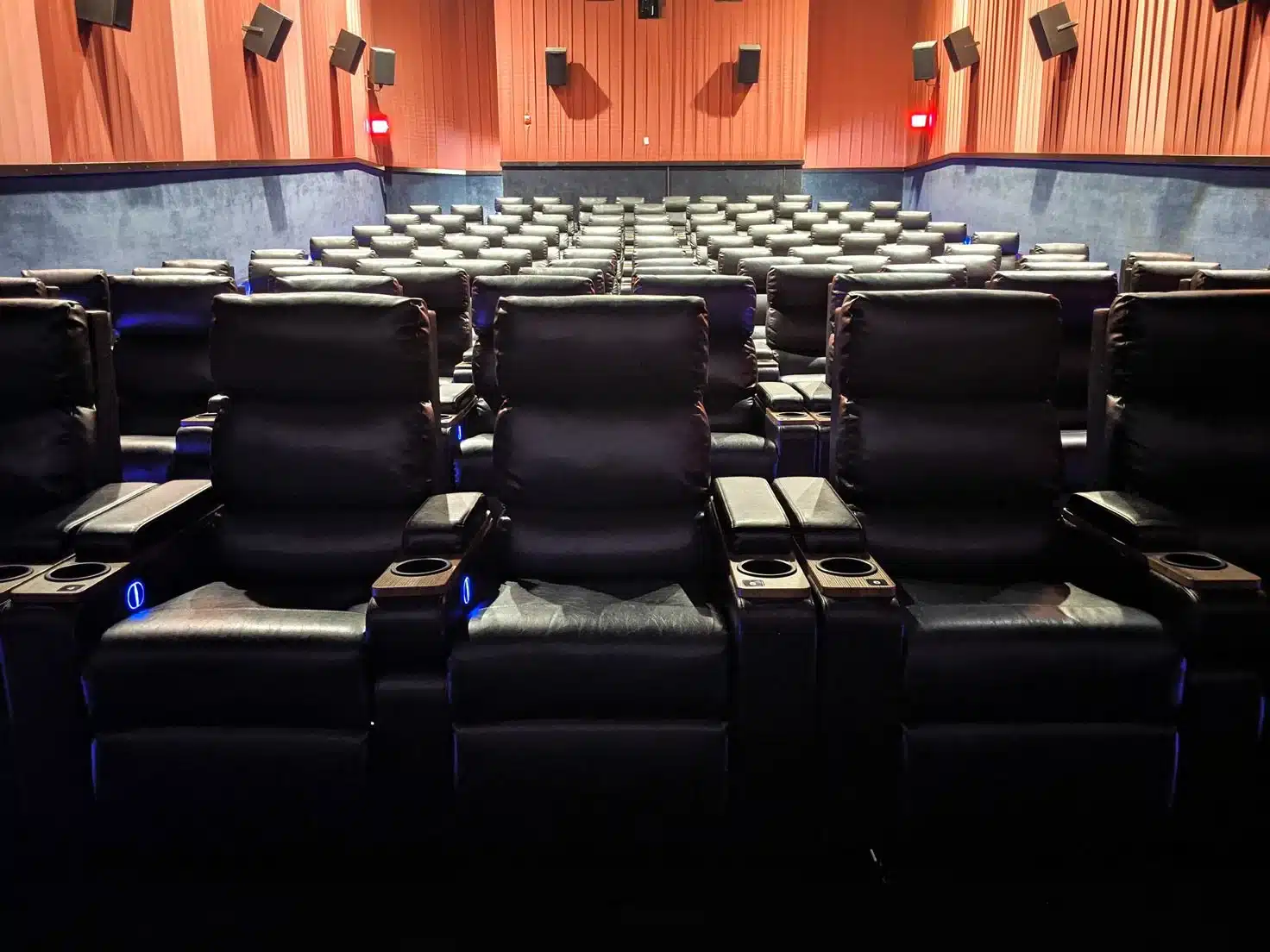 a Frederick Movie Theatre Featuring Rows of Empty Black Leather Recliner Seats Each with Cup Holders and Armrests Red Walls with Blue Lower Panels and Side mounted Speakers Complete the Dimly Lit Room Facing the Screen