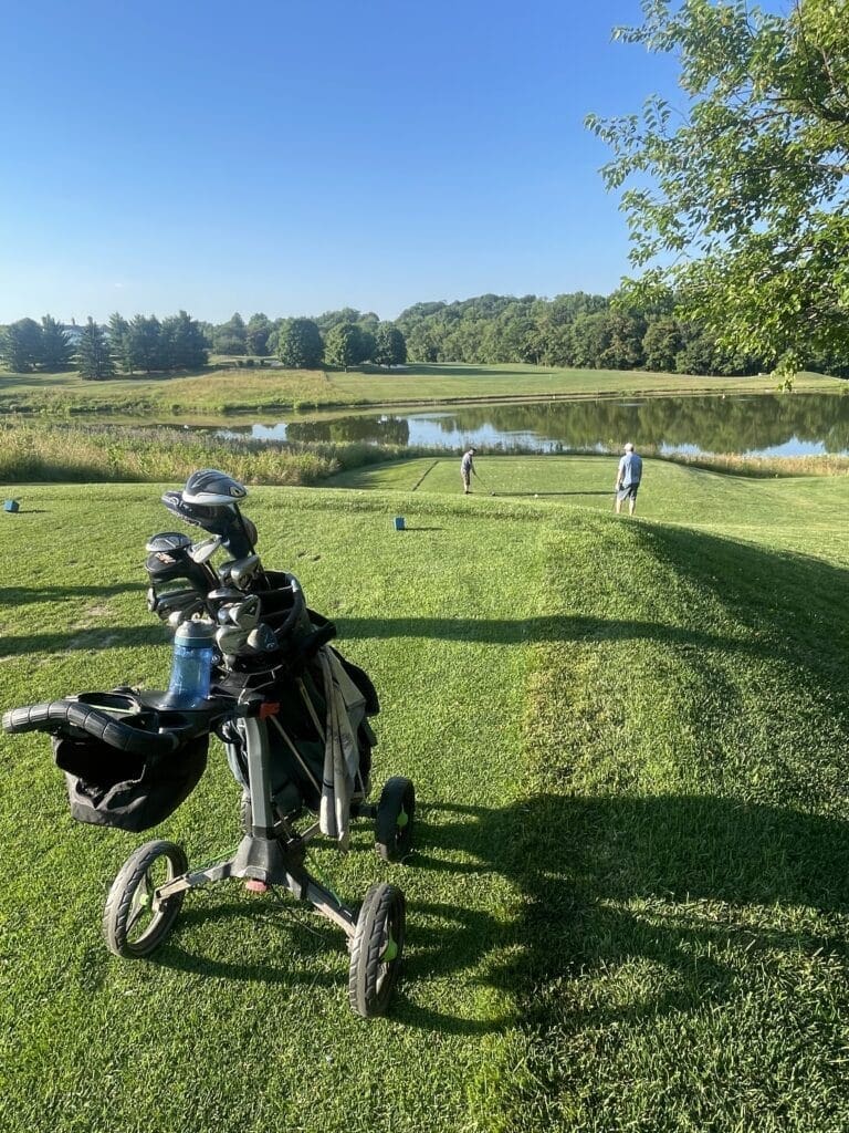 a Golf Bag on a Push Cart Sits on Green Grass in the Foreground Two Golfers Near a Tee by a Pond Surrounded by Trees and Under a Clear Blue Sky Enjoy a Sunny Day at the Mt Airy Md Course