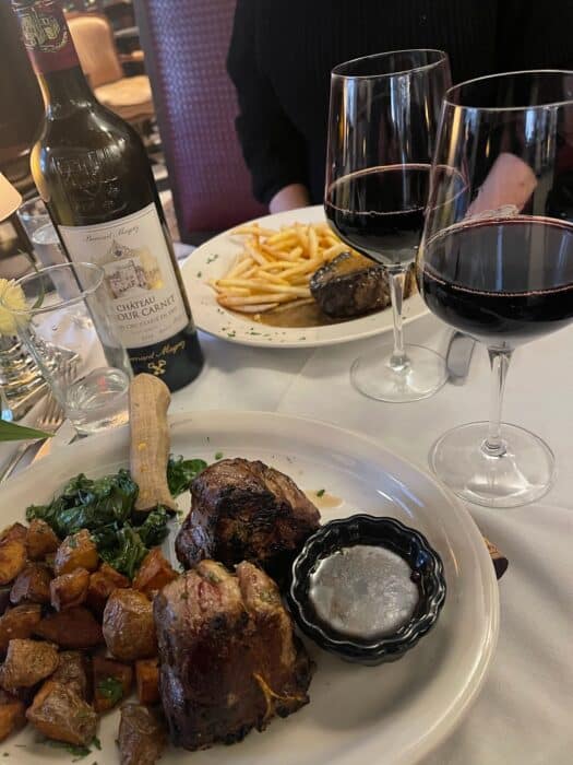 Two Plates of Food and Two Glasses of Red Wine Are on a Table at One of the Best Steakhouses Frederick Md Has to Offer One Plate Features Roasted Potatoes Greens and Meat the Other Has Steak and French Fries a Bottle of Château Moulin Wine is Nearby