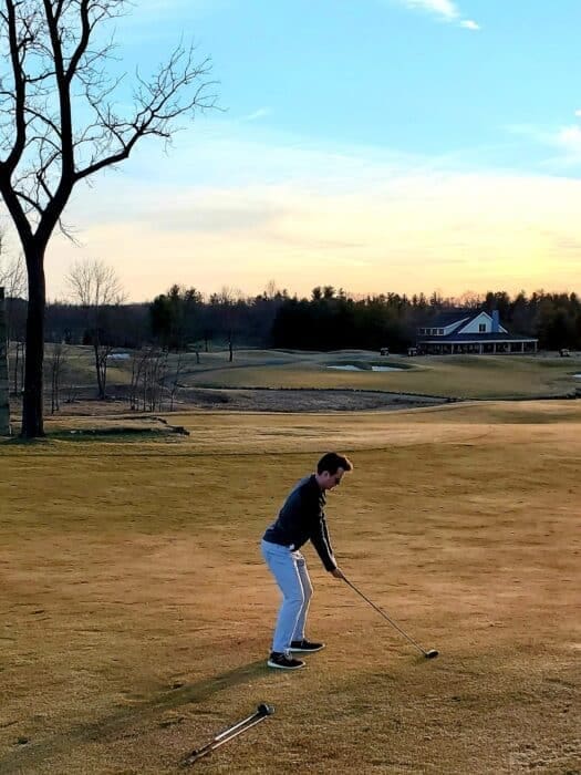 a Person Stands on a Golf Course in Md in the Late Afternoon Preparing to Swing a Club the Sky is Partly Cloudy with Orange and Blue Hues a Large Tree is Left and a Clubhouse with a Porch is Visiblelike Golf Courses Near Frederick
