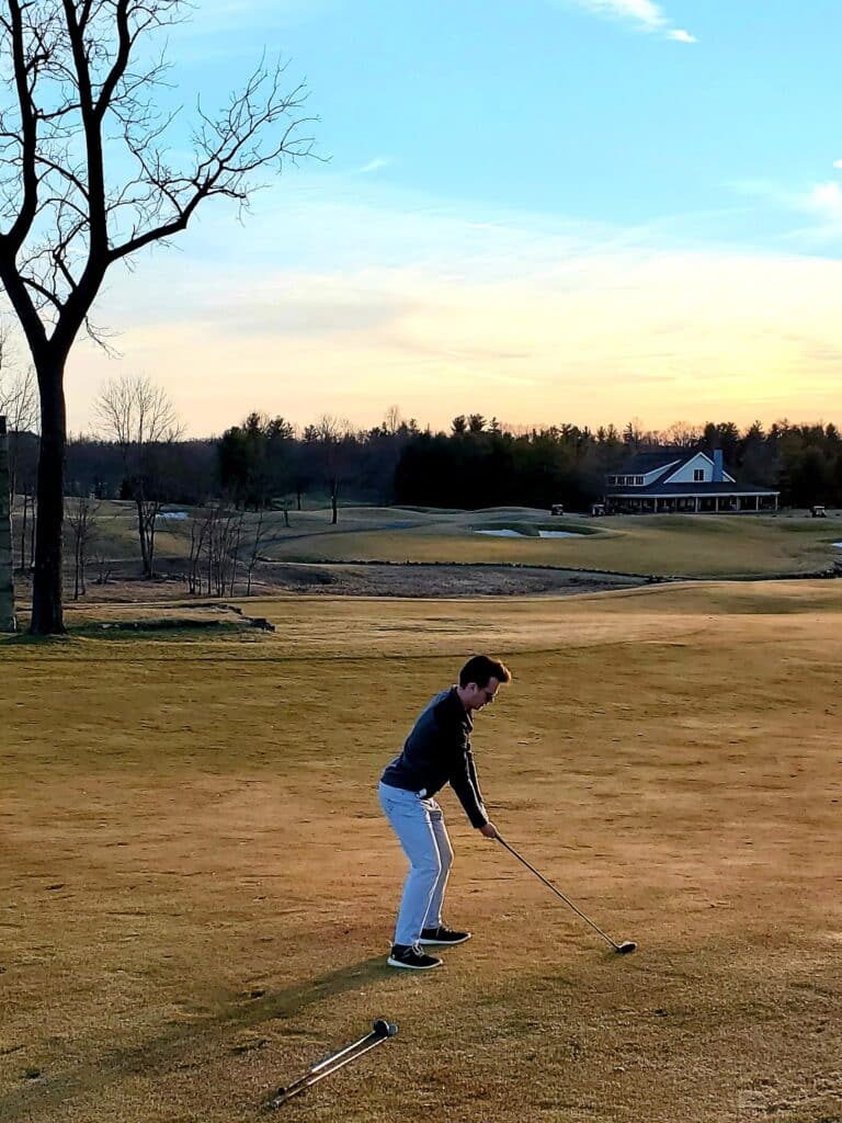 a Person Stands on a Golf Course in Md in the Late Afternoon Preparing to Swing a Club the Sky is Partly Cloudy with Orange and Blue Hues a Large Tree is Left and a Clubhouse with a Porch is Visiblelike Golf Courses Near Frederick