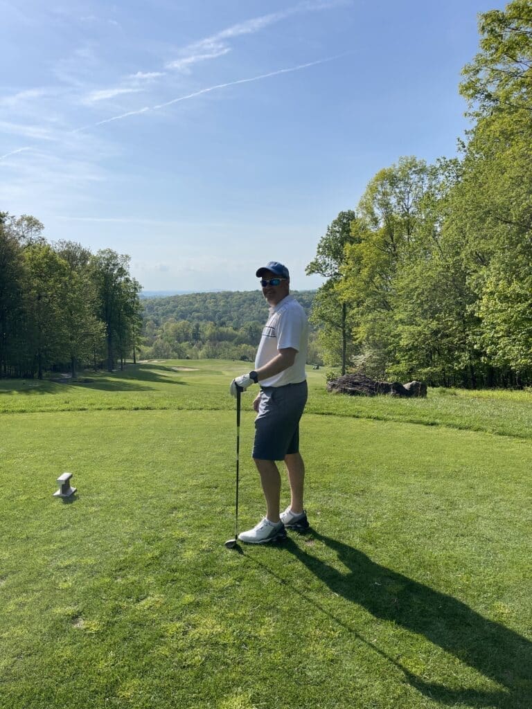 Whiskey creek golf course hole 5 a Man in a White Shirt Gray Shorts White Shoes and a Cap Stands on a Sunny Golf Course Near Frederick Md Holding a Golf Club He Faces the Camera and Smiles Surrounded by Green Grass Trees and Distant Hills Under a Clear Blue Sky