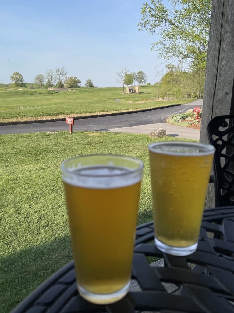Two Glasses of Light Beer Sit on a Black Metal Table Outdoors Overlooking Green Grass and Trees at a Golf Course in Md the Clear Blue Sky and Paved Path Create a Relaxing Scene Perfect for Enjoying Golf Courses Near Frederick