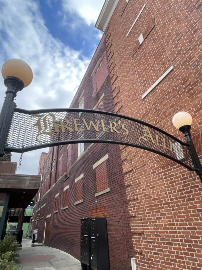 a Decorative Black Iron Archway with Gold Letters Reading Brewers Alley One of the Best Bars in Frederick Stands Between Two Lampposts in Front of a Tall Red Brick Building the Sky is Partly Cloudy and a Sidewalk Runs Alongside the Building