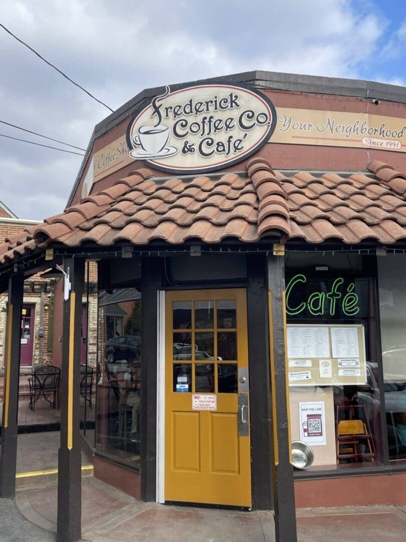 a Coffee Shop with a Brown Tile Roof Yellow Door Large Windows and a Sign Reading frederick Coffee Co  Cafe the Neon cafe Sign Glows in a Window Outdoor Seating is Visible with Tables and Chairs Along the Brick Sidewalkan Inviting Spot Nestled in One of the Charming Neighborhoods in Frederick Md