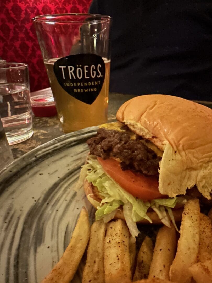 a Close Up of a Plate with the Best Burger Frederick Md Has to Offer Featuring Lettuce Tomato and Thick Cut Fries in the Background Sit a Glass of Light Beer with the Tröegs Independent Brewing Logo a Glass of Water and a Red Upholstered Seat