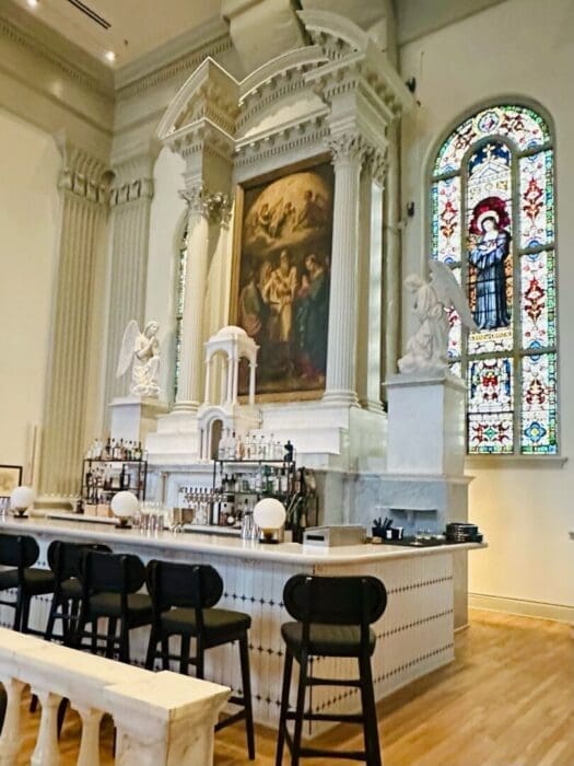 a Marble Bar with Black Chairs Sits Beneath a Large Religious Painting at Visitation Hotel Frederick Md Framed by White Columns and Two Angel Statues a Stained Glass Window of a Saint Blends Ornate Church Architecture with a Modern Bar Setup