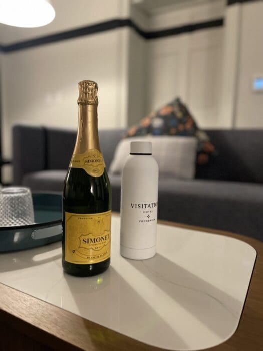 a Bottle of Simonet Sparkling Wine and a White Visitation Hotel Frederick Md Branded Water Bottle Sit on a Modern Coffee Table in the Background a Sofa with Patterned Cushions and a Dimly Lit Stylish Living Room Setting Are Visible