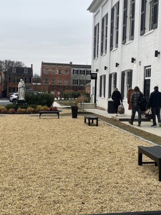 a Gravel Courtyard with Benches and a Planter Sits Beside the Visitation Hotel Frederick Md Featuring Tall Windows Three People Walk Toward the Entrance Carrying Bags Brick Buildings Shrubbery and a White Statue Appear in the Overcast Background