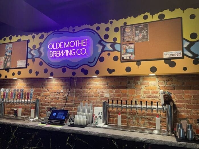 A bar counter with many beer taps sits below a brick wall. A colorful neon sign reads Olde Mother Brewing Co., one of the best breweries in Frederick MD. Flyers and corkboards hang nearby, with cups, a monitor, and sanitizer bottles on the counter.