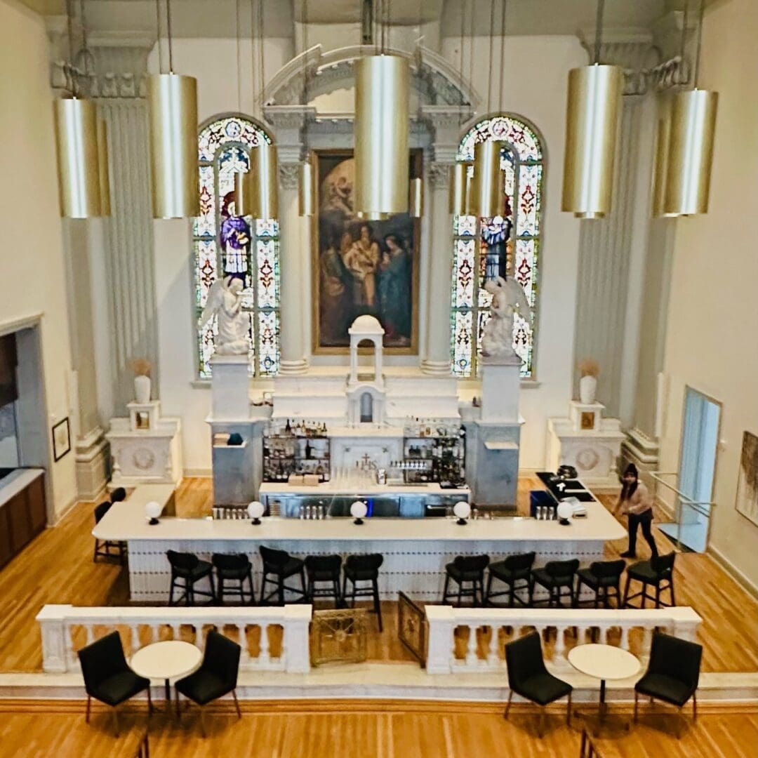 a Stylish Bar Set Inside a Former Church This Unique Gem Among Restaurants in Frederick Md Features High Stained glass Windows Neoclassical Columns Gold Pendant Lights a Marble Bar Lined with Bottles Black Barstools and Round Tables on a Wooden Floor
