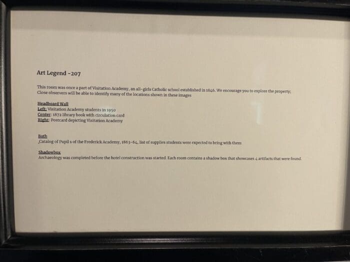 Framed Sign Titled Art Legend  207 Explains This Room at Visitation Hotel Frederick Md Was Once Part of Visitation Academy an All Girls Catholic School It Highlights a Headboard Wall Photo a Catalog of Student Belongings and Shadow Box Artifacts