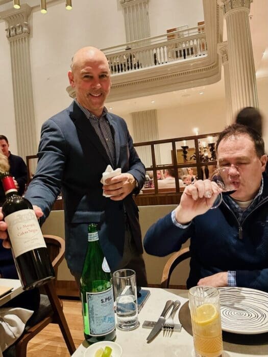 a Smiling Server in a Suit Presents a Bottle of Red Wine to a Guest at the Best Steakhouse Frederick Md Where Tall White Columns Frame the Restaurant the Table Features San Pellegrino a Cocktail and a Plate with a Napkin