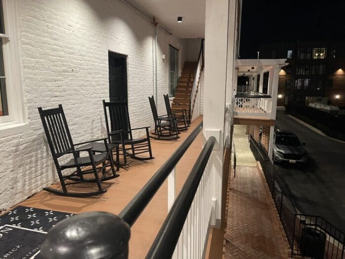 a Nighttime View of an Outdoor Hallway at Visitation Hotel Frederick Md Features Wooden Floors White Brick Walls Black Doors and Four Rocking Chairs Stairs Lead Upward Railings Line the Walkway and a Car is Parked Below Near Brick Paving