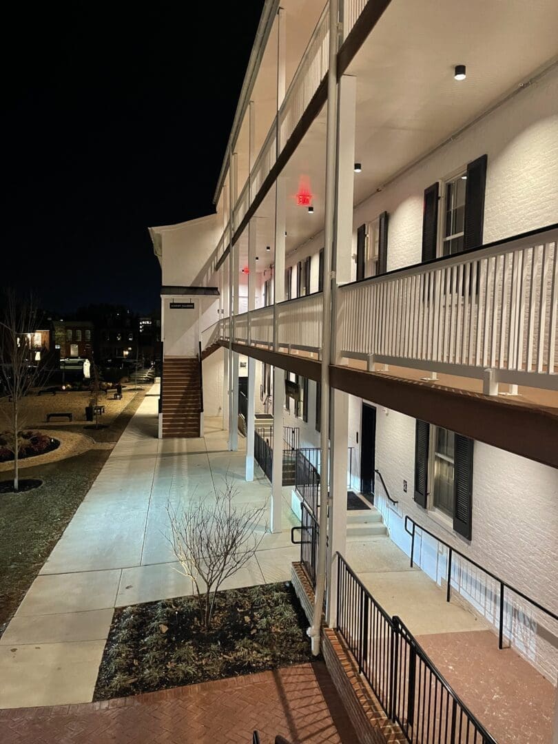 a Two story White Brick Building at Night Features a Lit Walkway Black Shutters on Windows and White Railings External Stairs Lead to the Upper Floor Leafless Trees and Landscaped Areas Border the Walkway at Visitation Hotel Frederick Md