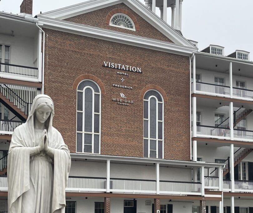 a White Statue of the Virgin Mary Stands with Hands in Prayer Before a Brick Building Labeled Visitation Hotel Frederick in Frederick Maryland the Building Has Tall Arched Windows White Trim and a Small Rooftop Tower Under an Overcast Sky