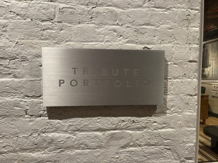 a Brushed Metal Sign with the Words Tribute Portfolio in Uppercase Letters is Mounted on a White Painted Brick Wall at Visitation Hotel Frederick Md Featuring a Modern Minimalist Style and Casting a Slight Shadow on the Textured Surface