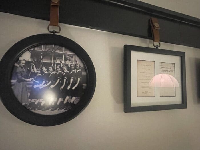 a Round Photo in a Black Frame Shows Women in Uniform Possibly from Visitation Hotel Frederick Md Standing Outdoors with an Older Woman Next to It a Rectangular Frame Displays Vintage Documents Both Hang from Brown Leather Straps on a Dark Rail